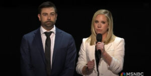 Two individuals standing side by side on an MSNBC broadcast. The woman on the right holds a microphone and appears to be speaking, wearing a white jacket over a dark top. The man on the left wears a dark blue suit with a white shirt and black tie, looking attentively forward. The background is dark, focusing attention on the speakers.